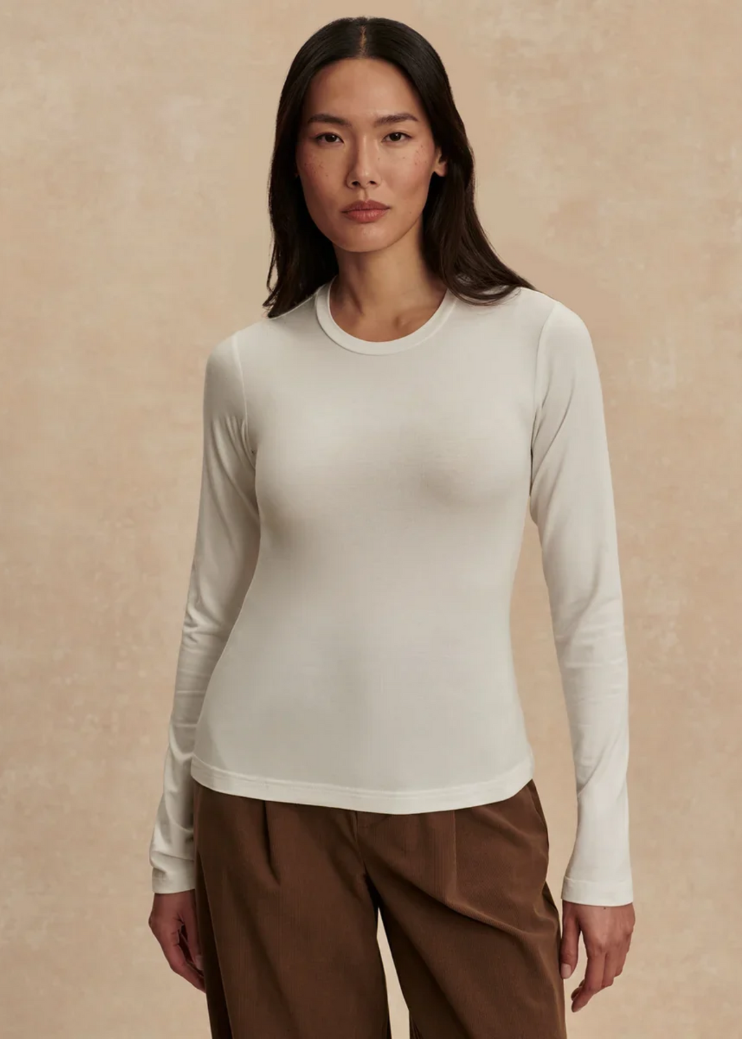 Woman wearing a white fitted long sleeve tee against a tan background.