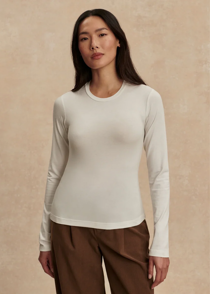 A woman wearing a fitted, long-sleeve crewneck top in off-white/cream, paired with brown pants, standing against a textured beige background.