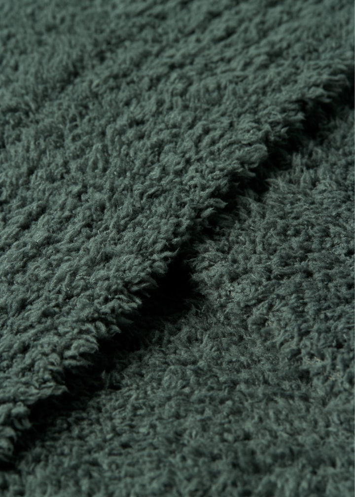 A close-up of a dark green, plush, textured fabric with a soft, looped pile and a folded edge showing the depth of the material.