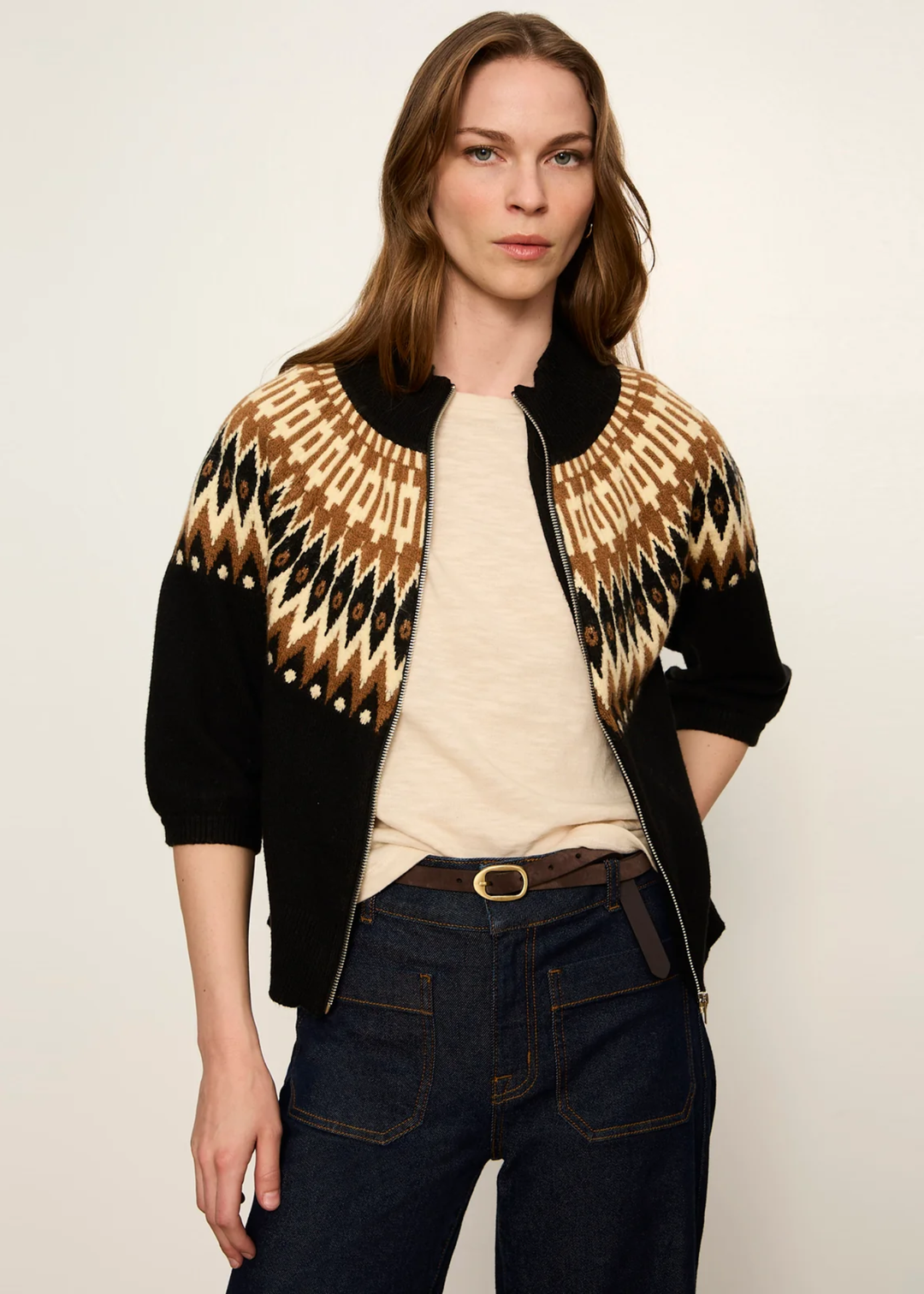 A woman modeling a black knit zip-up cardigan with a tan and black geometric pattern on the shoulders and upper chest, worn over a cream top and dark blue jeans against a plain light background.