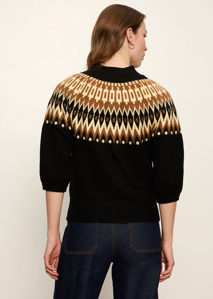 A woman shown from the back wearing a black knit sweater with a geometric Fair Isle pattern in beige, brown, and black across the shoulders and upper back, paired with dark blue jeans against a plain off-white background.