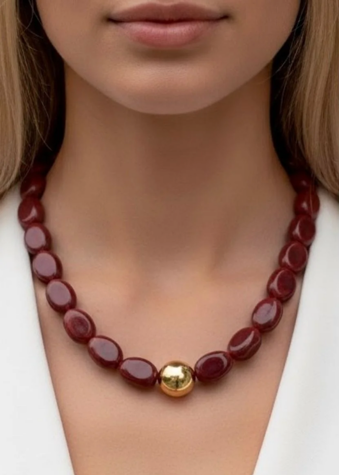 A close-up of a model wearing a dark red beaded necklace with a polished gold central bead, styled with a white top.