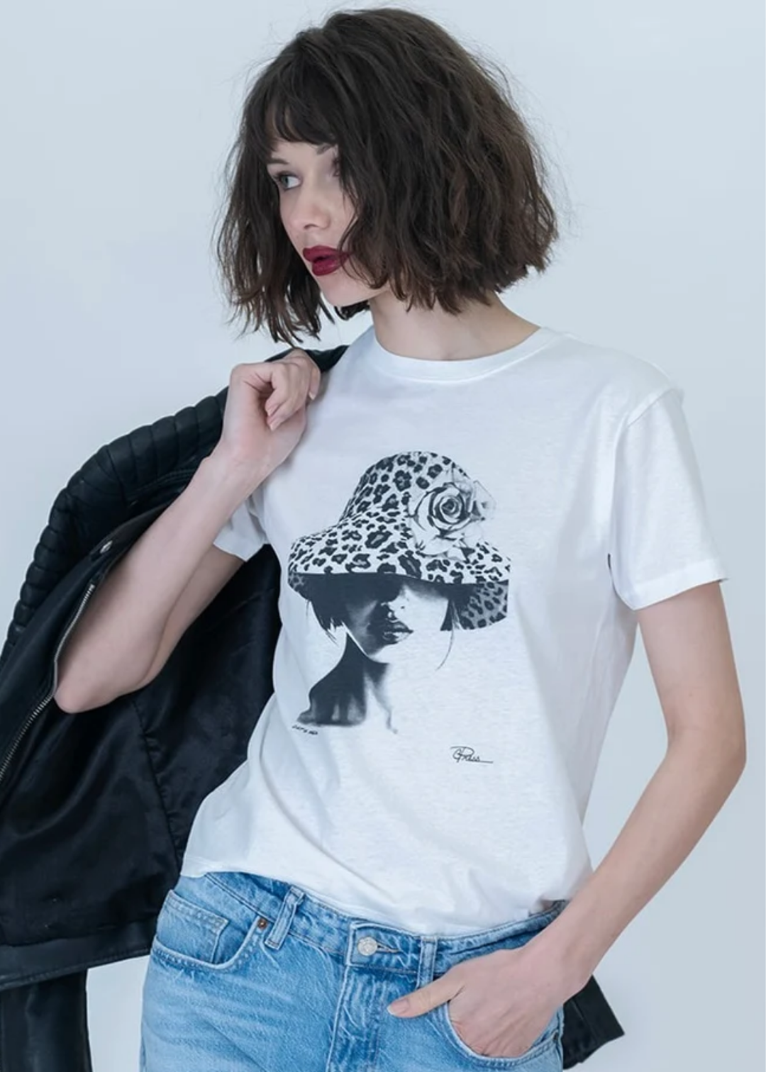 A woman wearing a white graphic t-shirt with a black and leopard-print illustration, styled with blue jeans and a black jacket draped over her shoulder, against a plain light gray background.