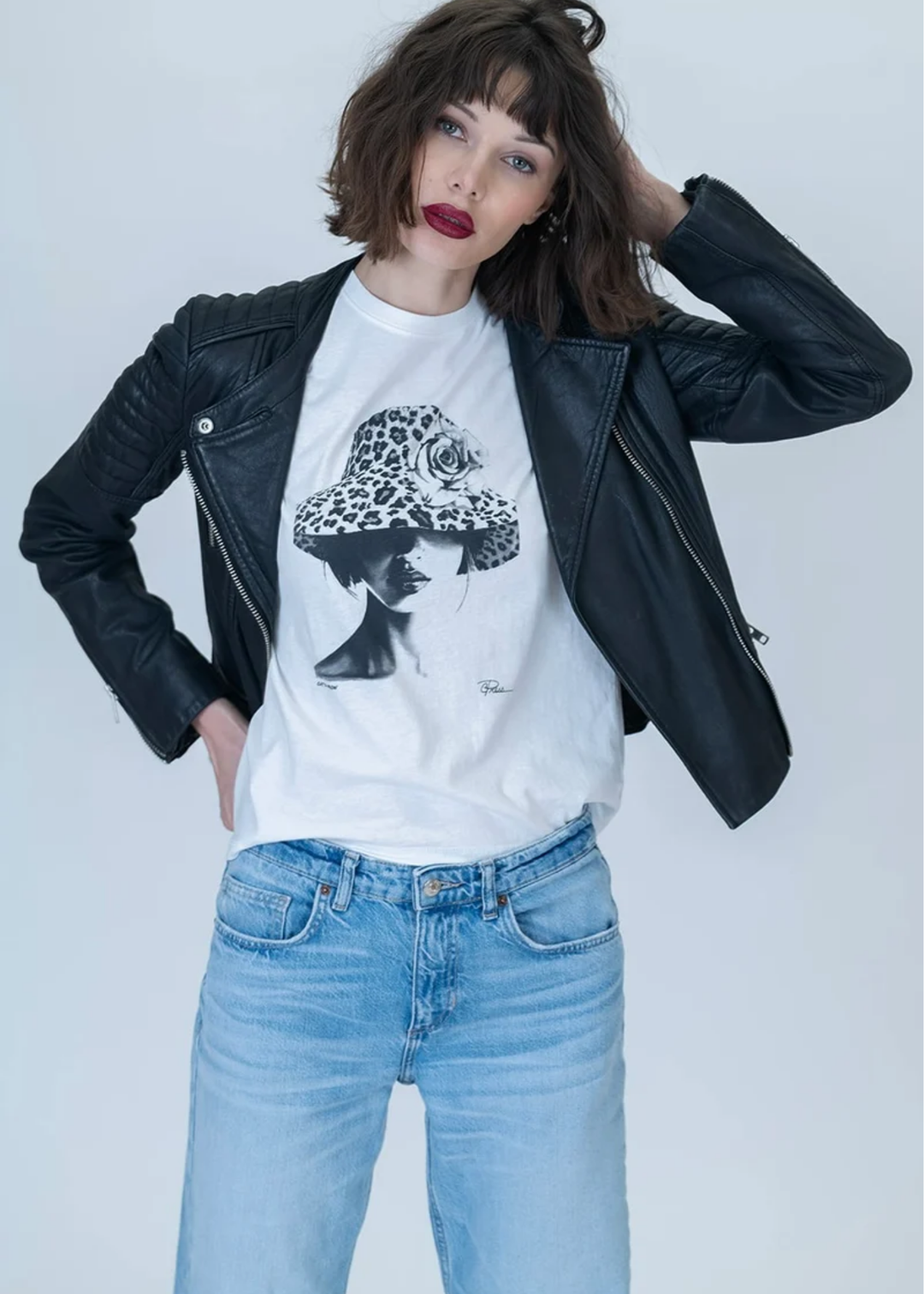 A model wearing a black leather jacket over a white graphic t-shirt with a black and white illustration of a woman in a hat, paired with light blue jeans, against a light gray studio background.