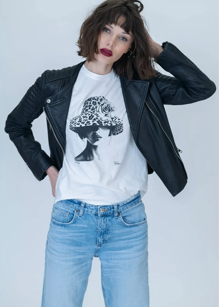 A model wearing a black leather jacket over a white graphic t-shirt with a black and white illustration of a woman in a hat, paired with light blue jeans, against a light gray studio background.