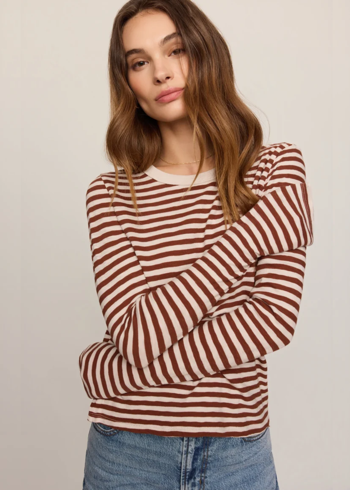 A woman wearing a rust brown and white horizontally striped long-sleeve knit top, paired with blue jeans, against a plain light beige background.