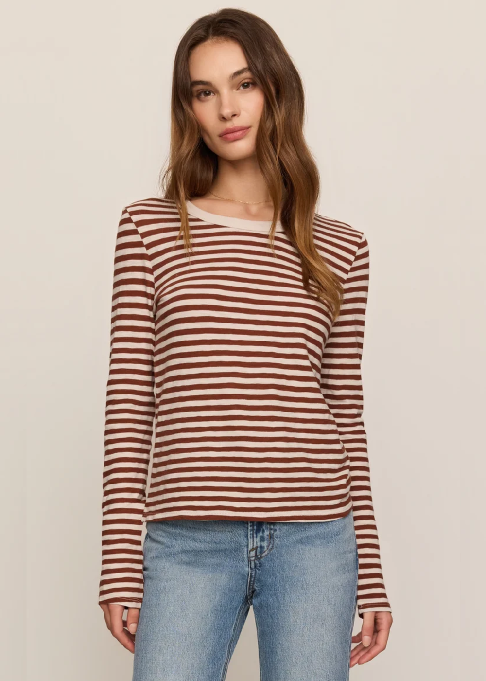 A woman modeling a long-sleeve knit top with horizontal rust-brown and beige stripes, paired with light-wash blue jeans, against a plain light beige studio background.