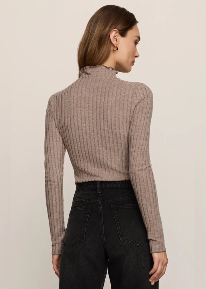 A woman shown from the back wearing a ribbed long-sleeve mock neck top in taupe and black jeans, against a plain light beige studio background.