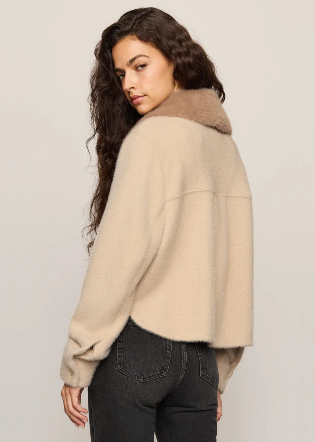 Woman wearing tan fuzzy jacket and facing backwards, and looking over her shoulder. 