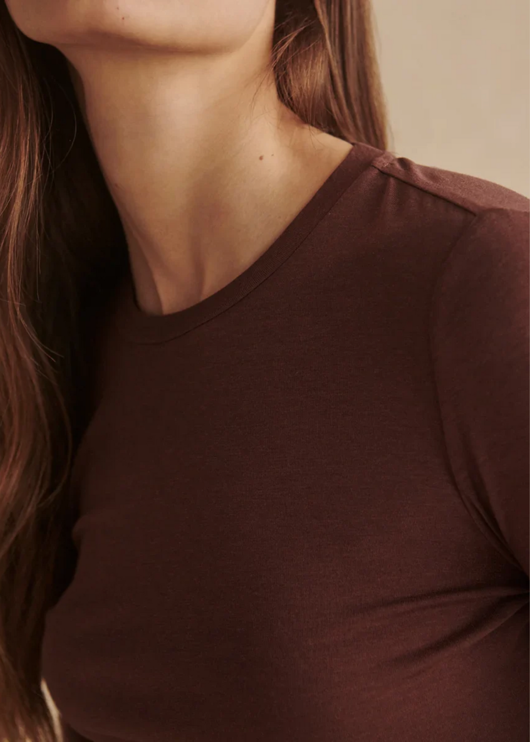 Close-up of a woman wearing a dark brown crew-neck long-sleeve top, showing the fabric texture and shoulder detail against a soft beige background.