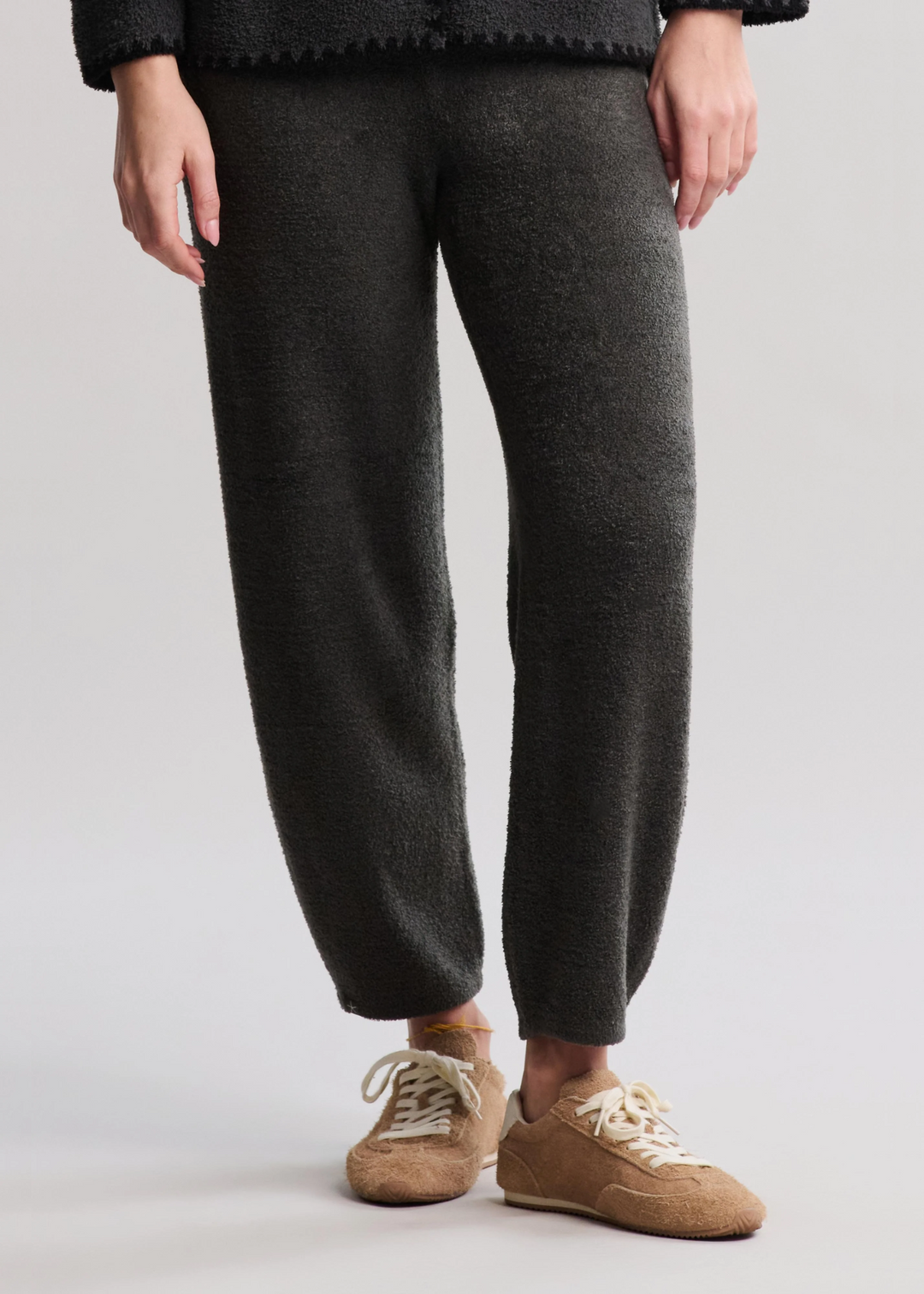 A woman wearing dark green/grey barrel pants with tan tennis shoes standing in front of a white background