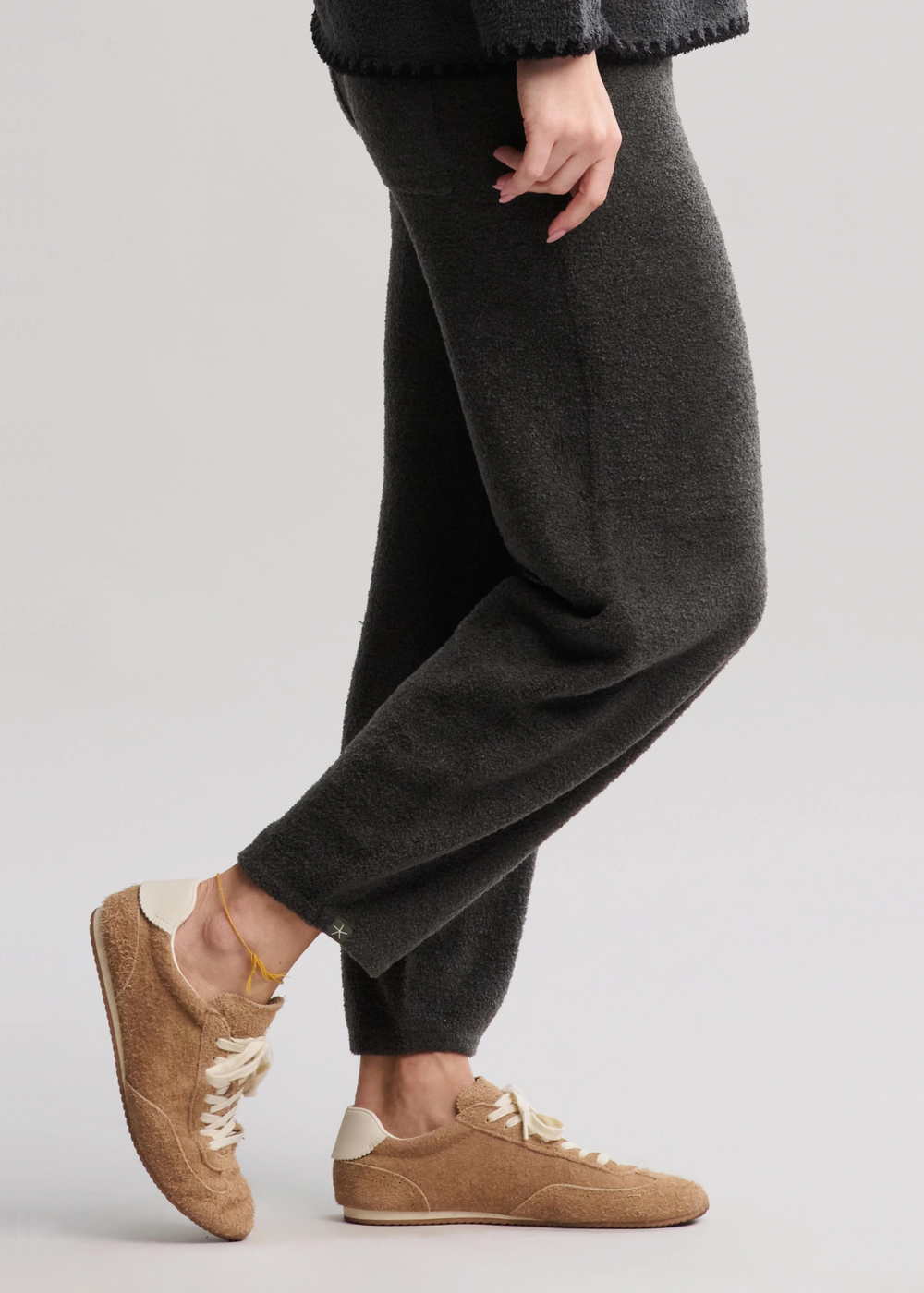 A woman turned to the side and wearing dark green/grey barrel pants with tan sneakers standing in front of a white background
