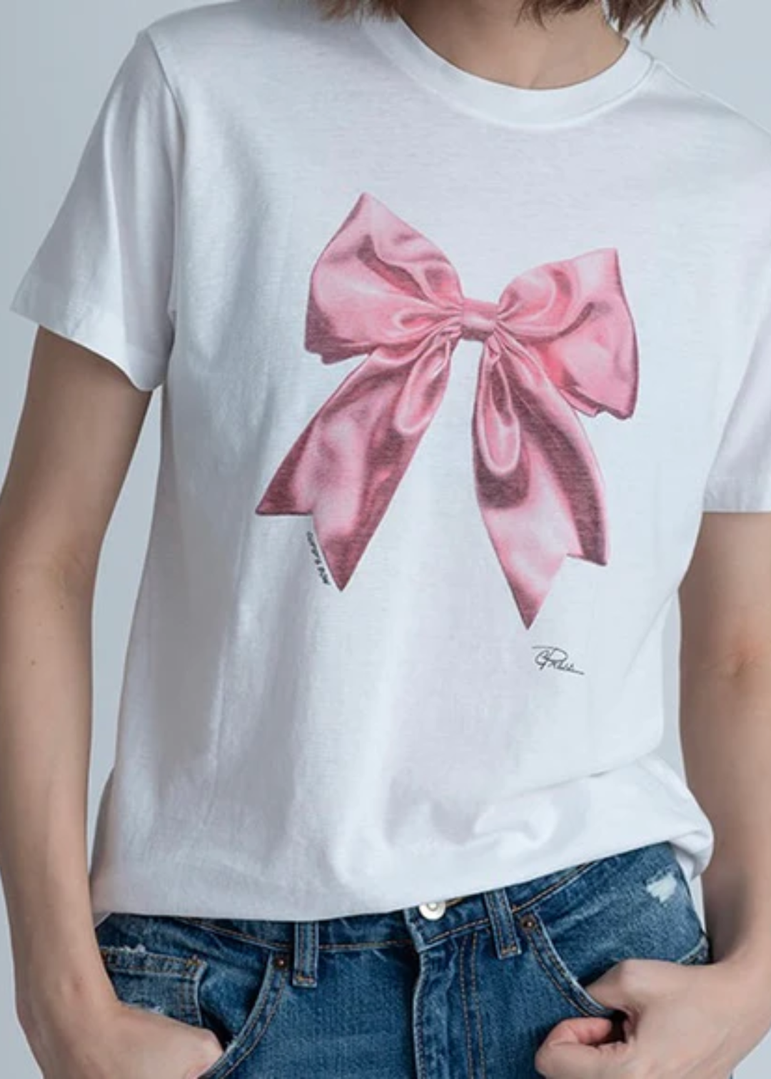 Woman wearing a white t-shirt with a pink bow graphic with jeans in front of a grey background