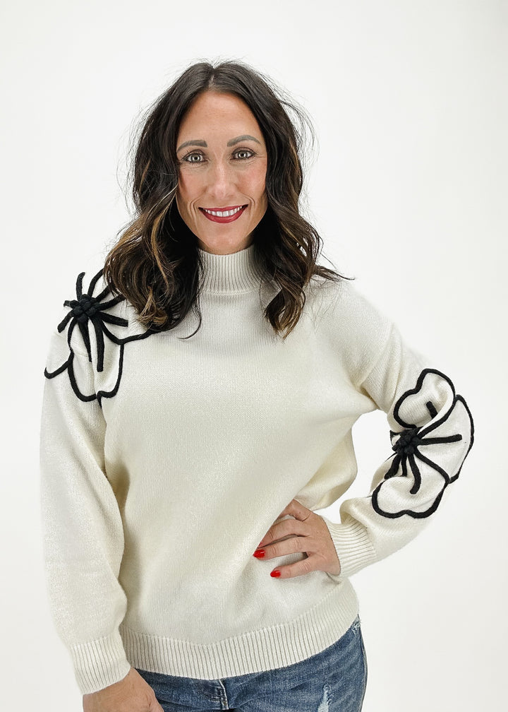 A woman wearing a cream sweater with black floral shoulder embellishments and long sleeves, styled with jeans for a chic and unique casual look.