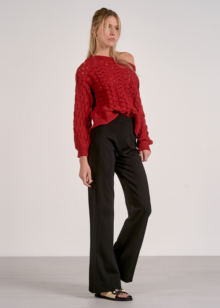 Woman wearing a red off the shoulder open knit sweater with black pants against a grey background