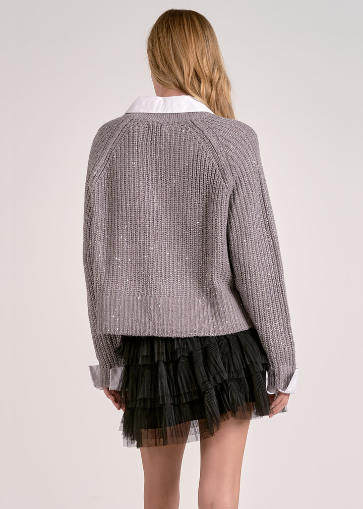 Woman turned around wearing a grey sparkly sweater layered over a white button down with a black mesh skirt