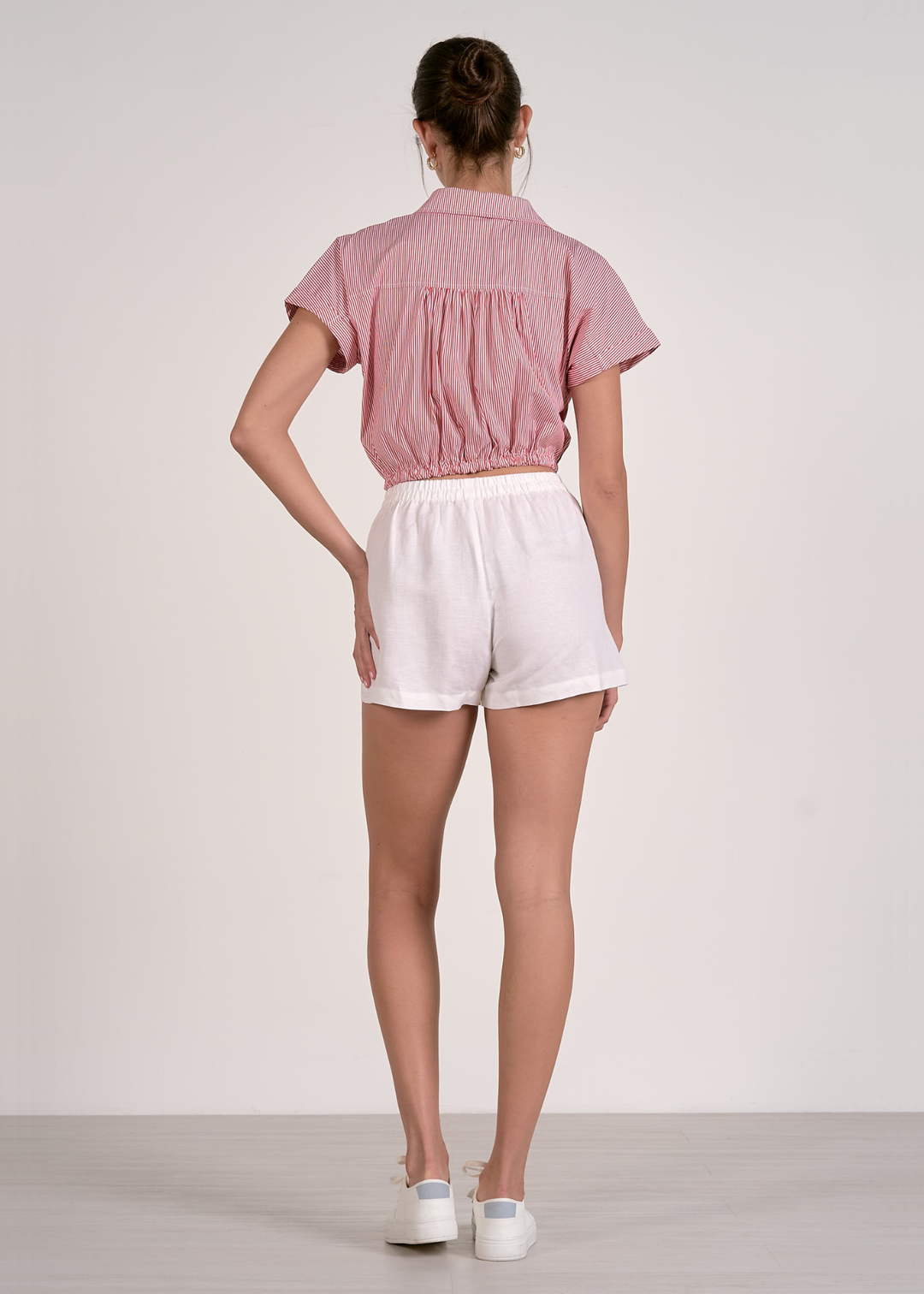 Woman turned around wearing a cropped and short sleeve red and white striped button down with white shorts against a grey background