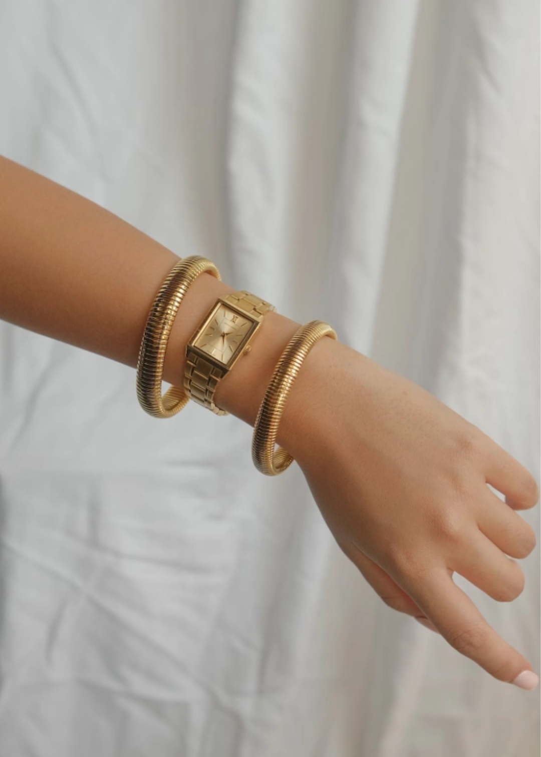 Woman wearing two gold bangle bracelets and a gold watch against a white background