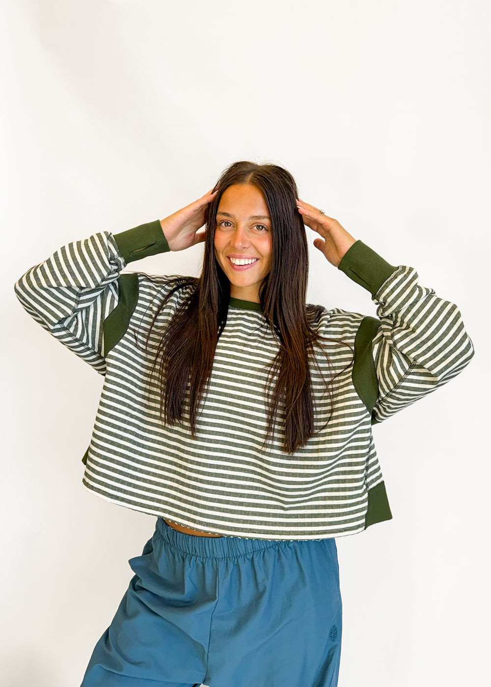 A woman posing in a cropped long-sleeve sweatshirt with green and white horizontal stripes and solid green ribbed cuffs and shoulders, paired with blue sweatpants against a plain white background.