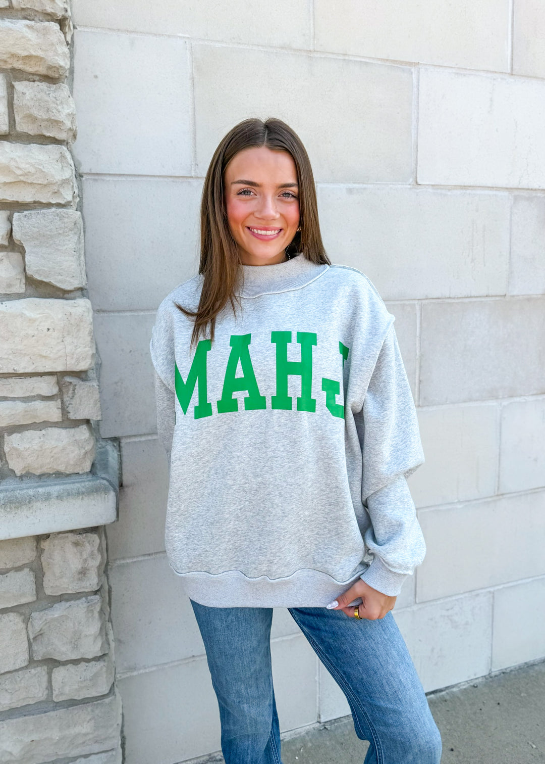 A woman wearing a light gray heather crewneck sweatshirt with 'MAHJ' printed in green varsity letters across the chest, paired with blue jeans, standing against a light stone wall.