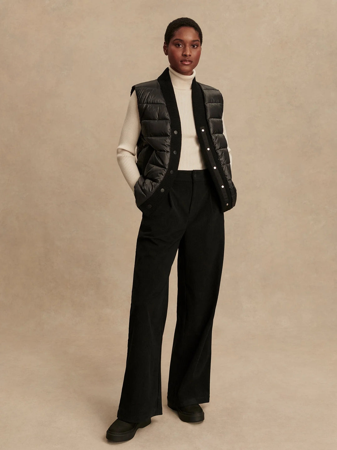 Woman wearing a black puffer vest over a white turtleneck and black pants in front of a tan background