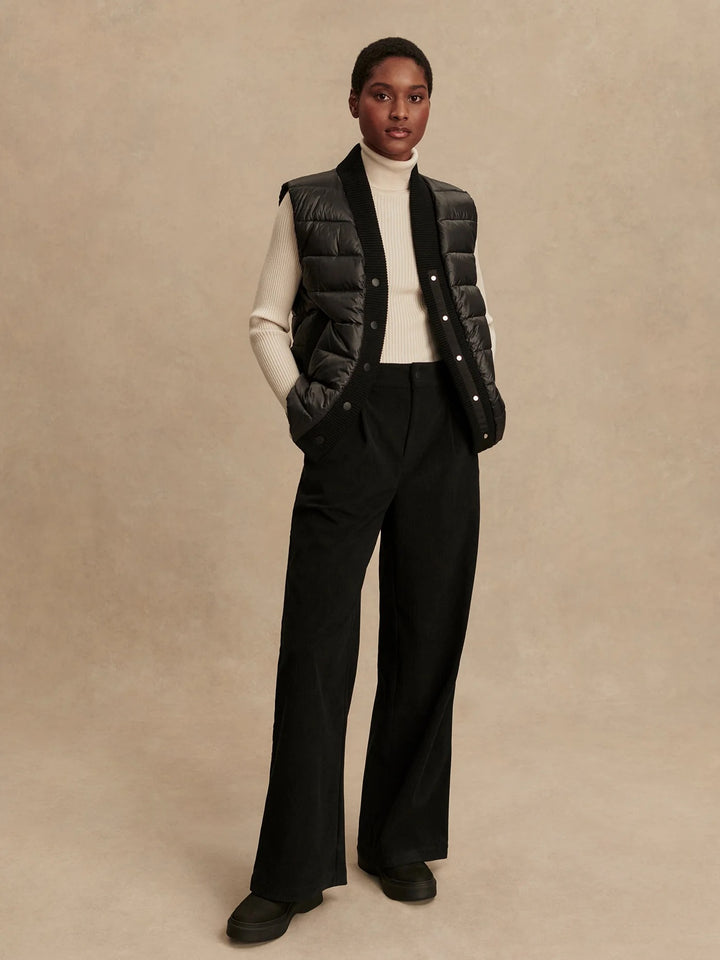 Woman wearing a black puffer vest over a white turtleneck and black pants in front of a tan background