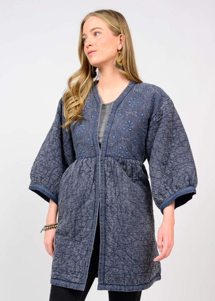 Woman wearing a long dark blue top with 3/4 length sleeves and a deep v-neck and floral embroidery standing in front of a white background