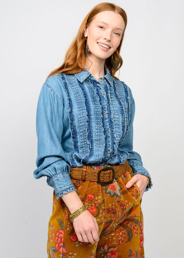 Woman wearing a denim ruffle button down top with colorful pants and hand in pocket in front of a white background