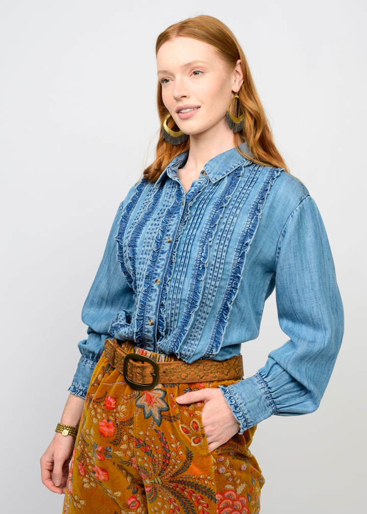Woman turned to the side wearing a denim ruffle button down top with colorful pants and hand in pocket in front of a white background