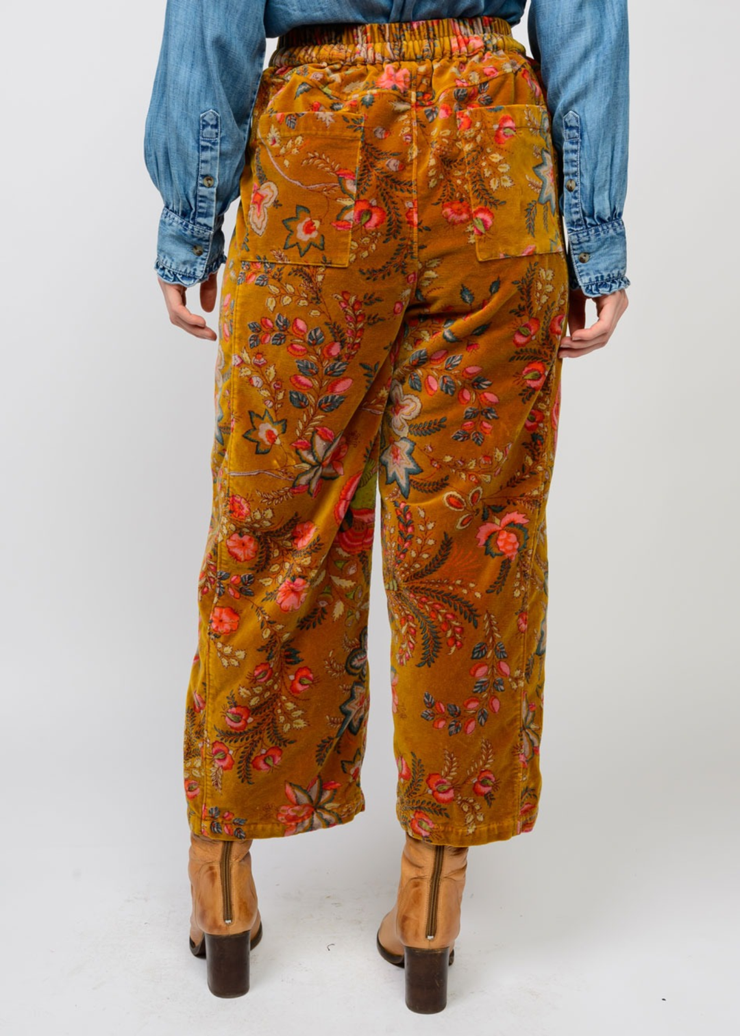 Woman turned around wearing gold barrel pants with a floral print and brown boots in front of a white background