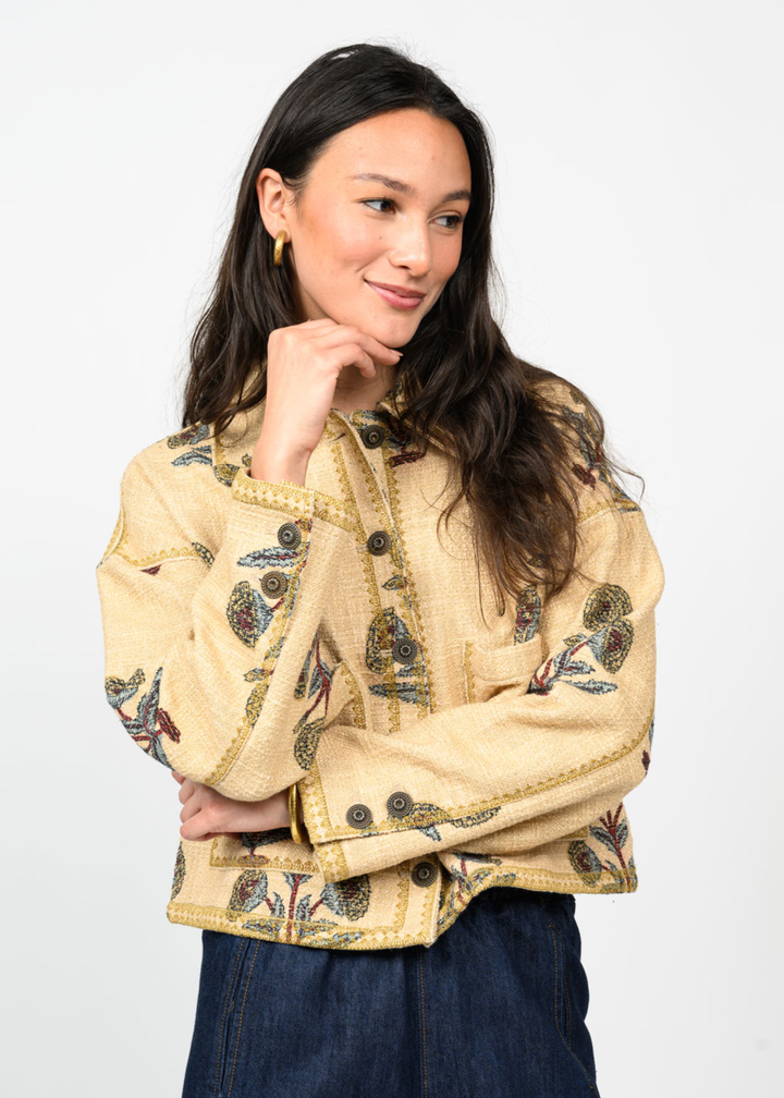 Woman with hand on chin wearing a tan jacket with a floral print and navy pants in front of a white background