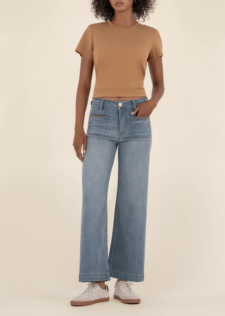 Woman wearing high rise, wide leg blue jeans with brown stitching on the front pockets and with a tan top in front of a tan background