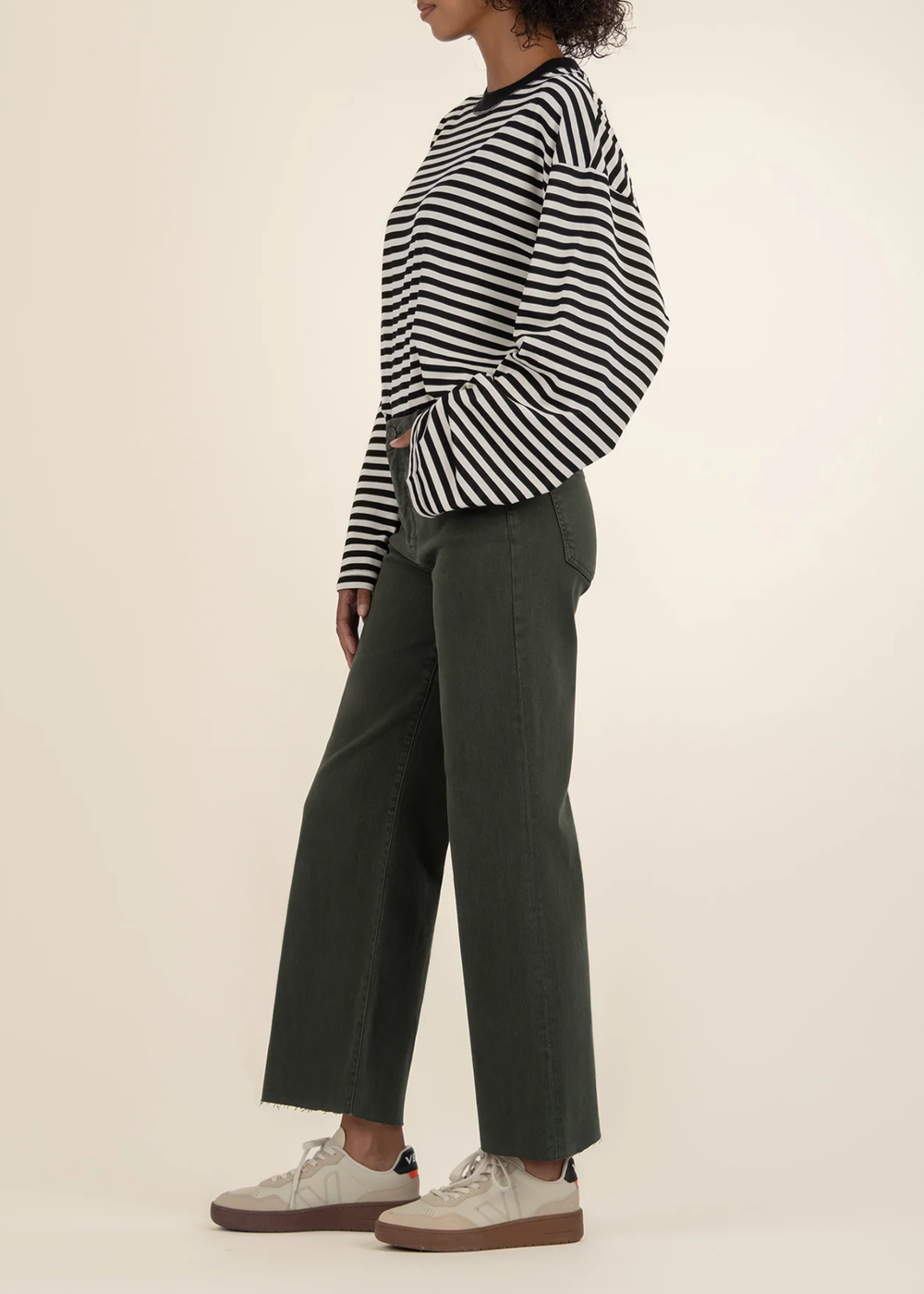 Woman turned to the side wearing dark green jeans with a black and white striped sweater and white sneakers in front of a white background