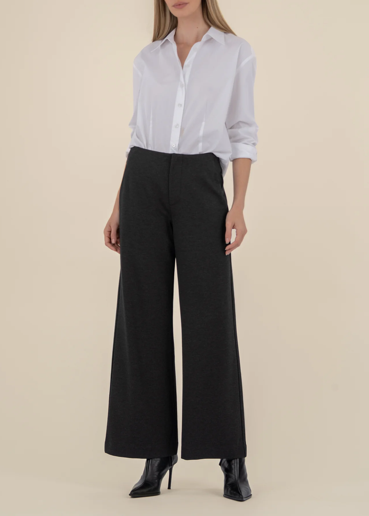 Woman wearing a white button down with charcoal grey wide leg pants and black heeled boots in front of a tan background