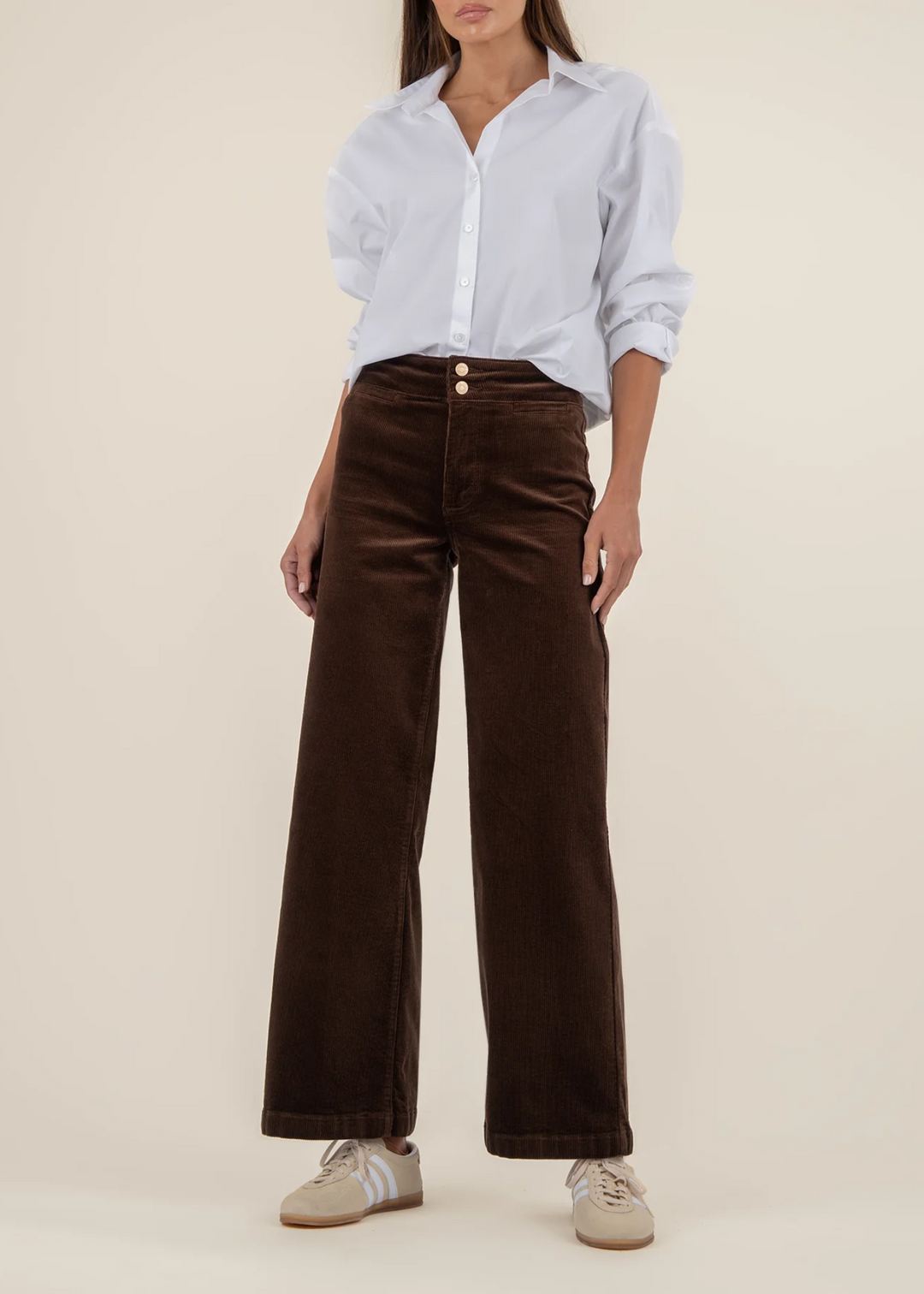 Woman wearing a white button down and dark brown corduroy wide leg pants with gold buttons in front of a tan background