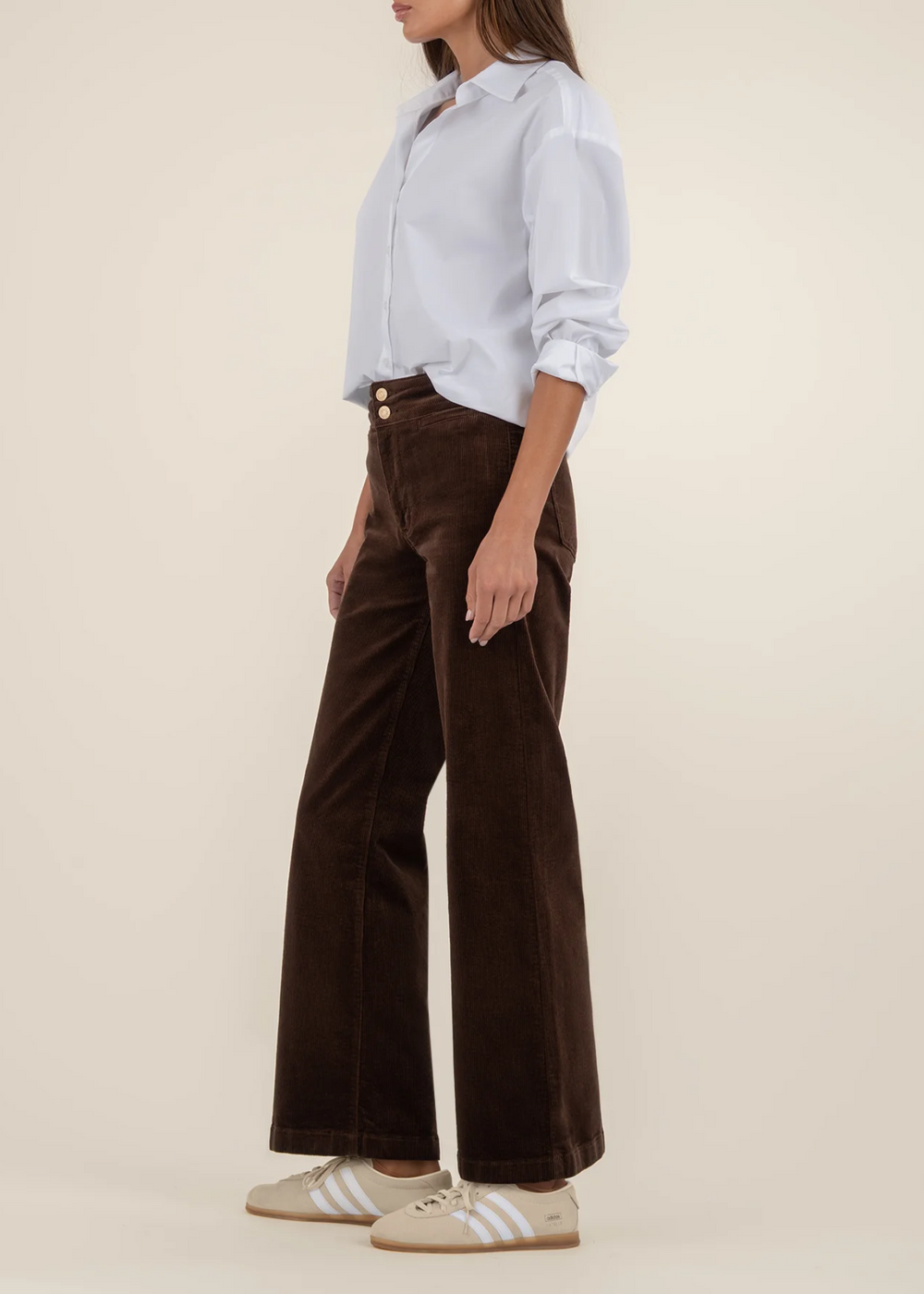 Side view of a woman wearing a white button down top with dark brown corduroy wide leg pants in front of a tan background