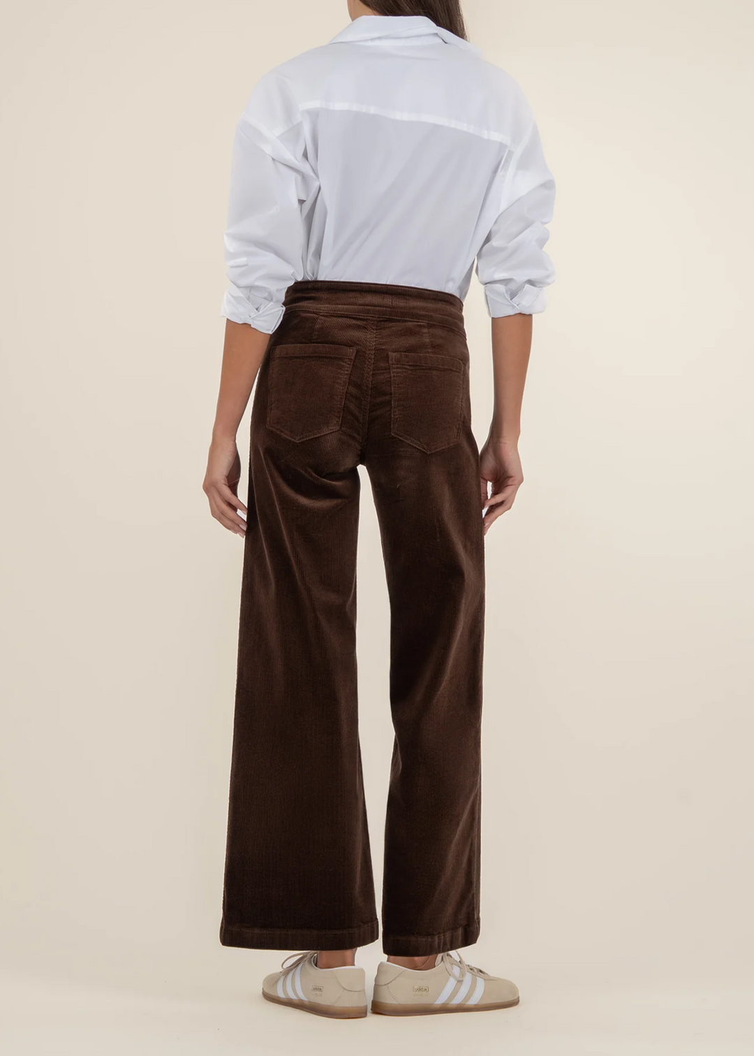 View from the back of a woman wearing a white button down top with dark brown corduroy wide leg pants in front of a tan background