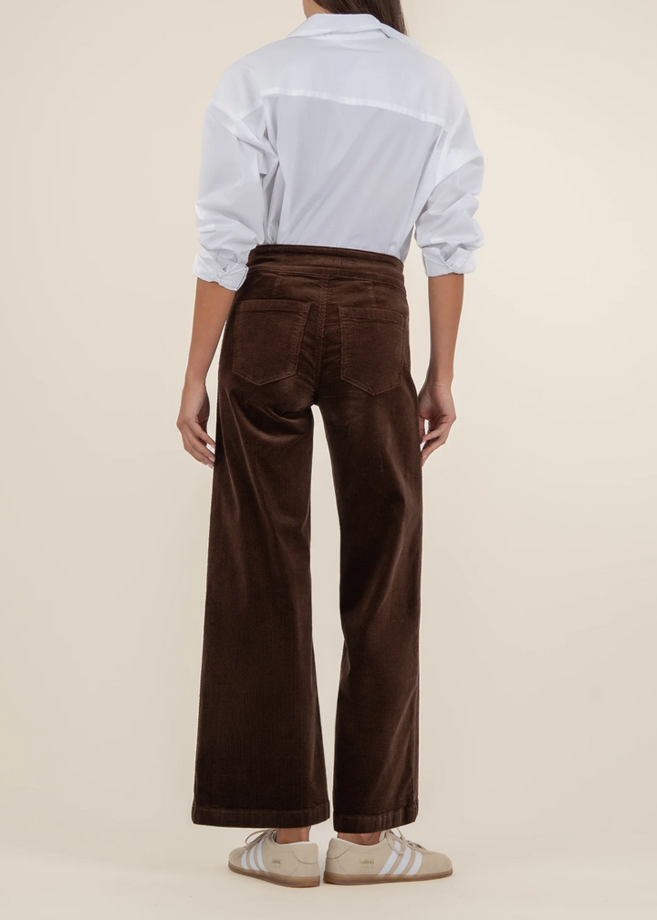 View from the back of a woman wearing a white button down top with dark brown corduroy wide leg pants in front of a tan background