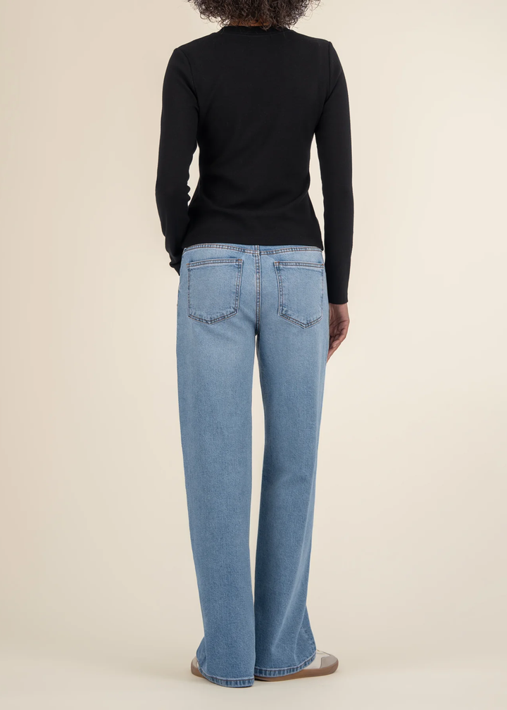 Woman turned around wearing a black long sleeve tee with jeans and white sneakers in front of a white background