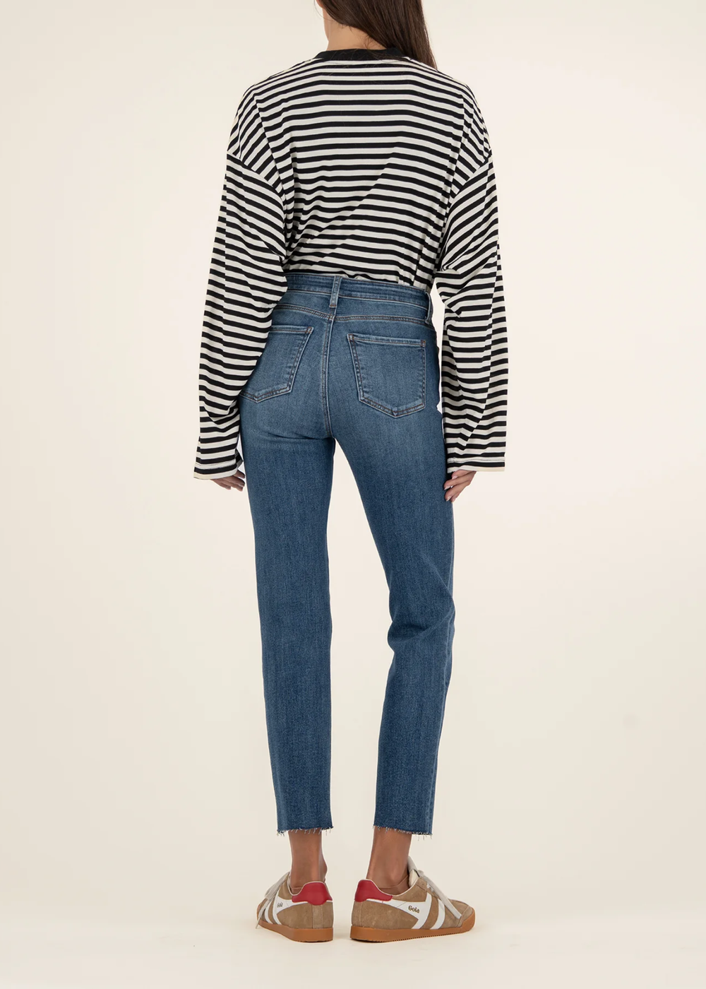 Woman turned around wearing medium wash blue ankle straight jeans with tan sneakers and a black and white striped sweater standing in front of a white background