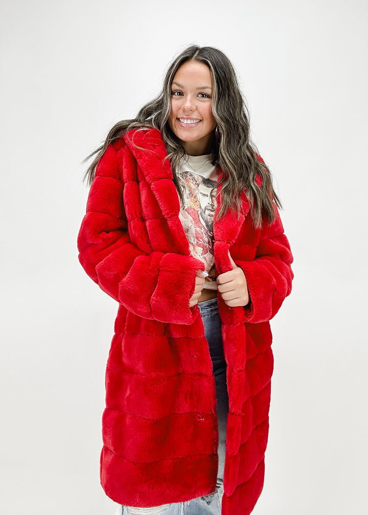 A woman wearing a long red faux fur coat with a plush texture, featuring a removable bottom and zip-off design for versatile styling.