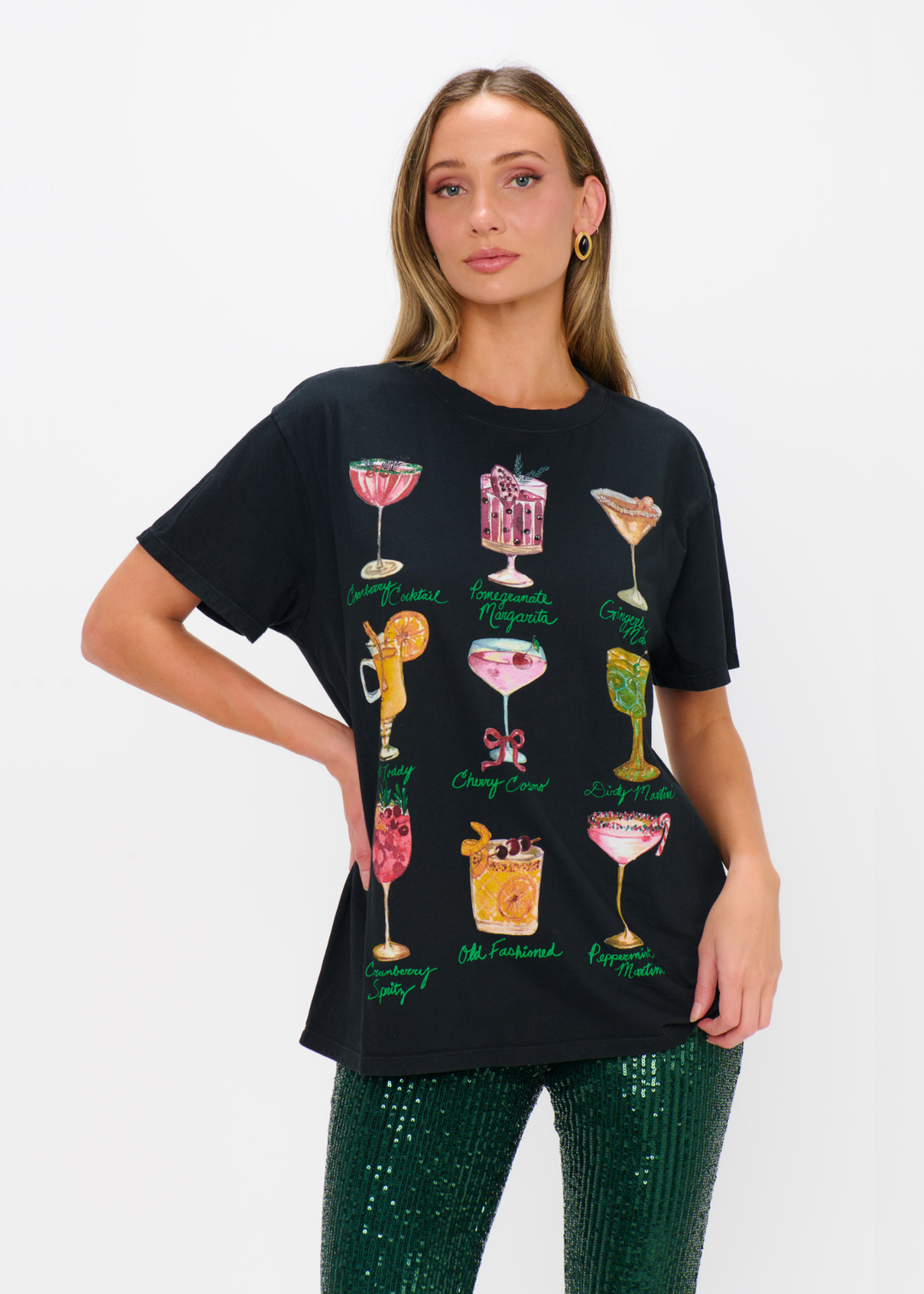Woman with a hand on hip wearing a black cocktails graphic tee with green sequin pants against a white background