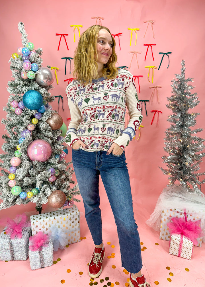 Person wearing a festive sweater standing between two decorated Christmas trees with a pink background.