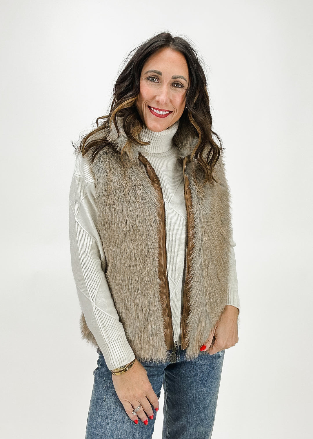 A woman wearing a reversible vest, featuring one side with faux fur and the other side a tan puffer design, styled with a white sweater and jeans for a versatile, cozy look.