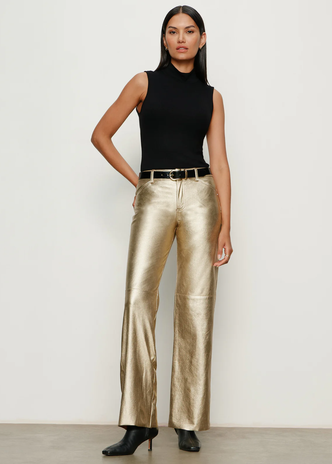 Woman wearing gold pants with black boots and a black top against a white wall