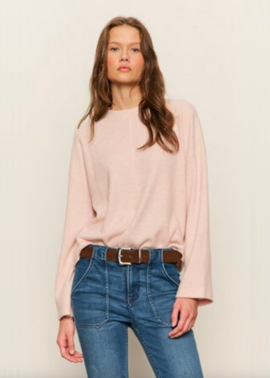 Woman wearing a light pink sweater with jeans and a brown belt standing in front of a grey background