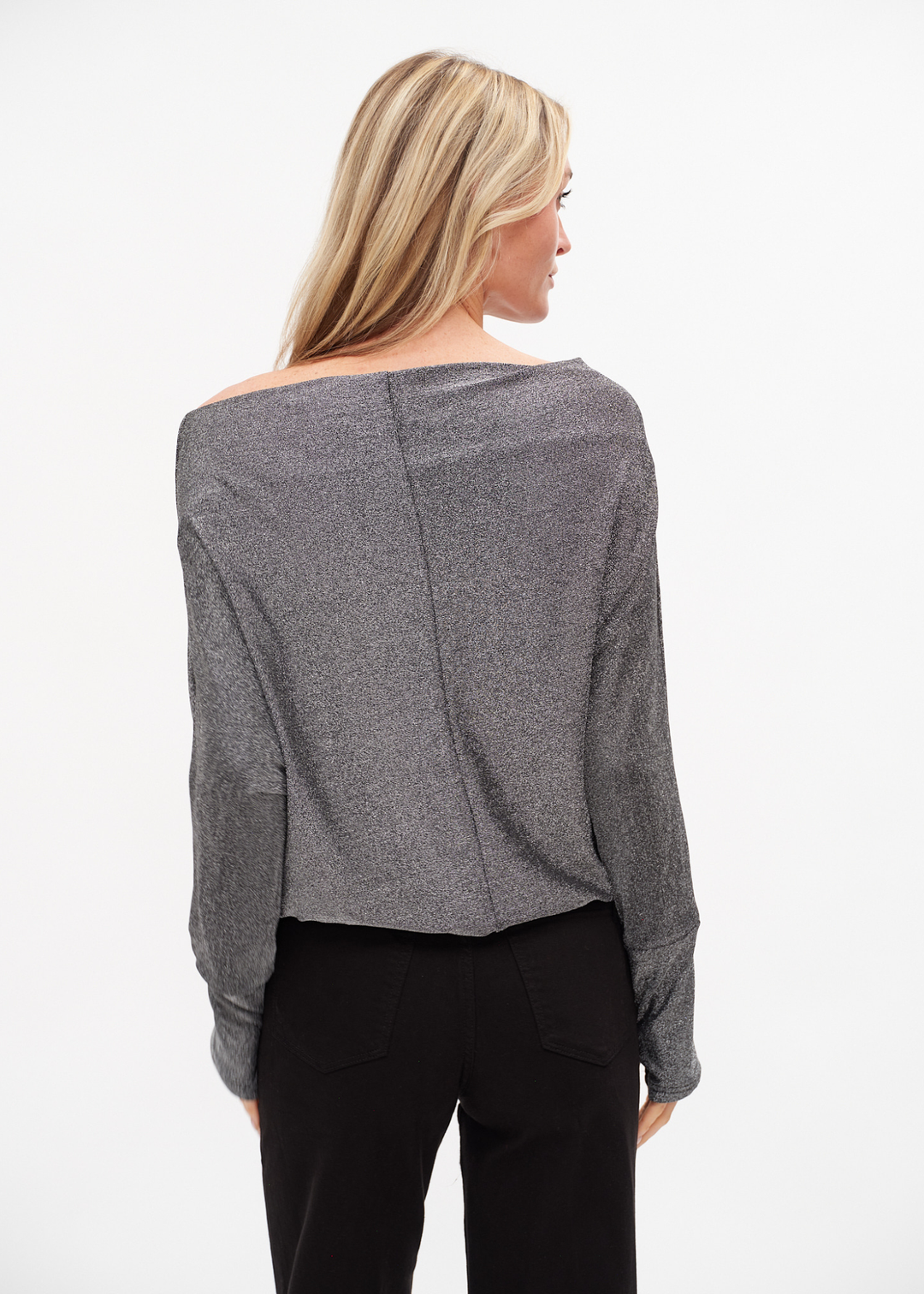 Woman turned around wearing a silver off the shoulder top with black jeans against a white background