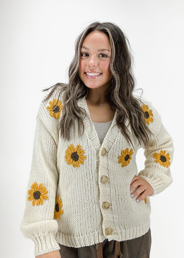 A woman wearing a chunky knit cream cardigan with embroidered sunflowers and a button closure, perfect for cozy fall and winter outfits.