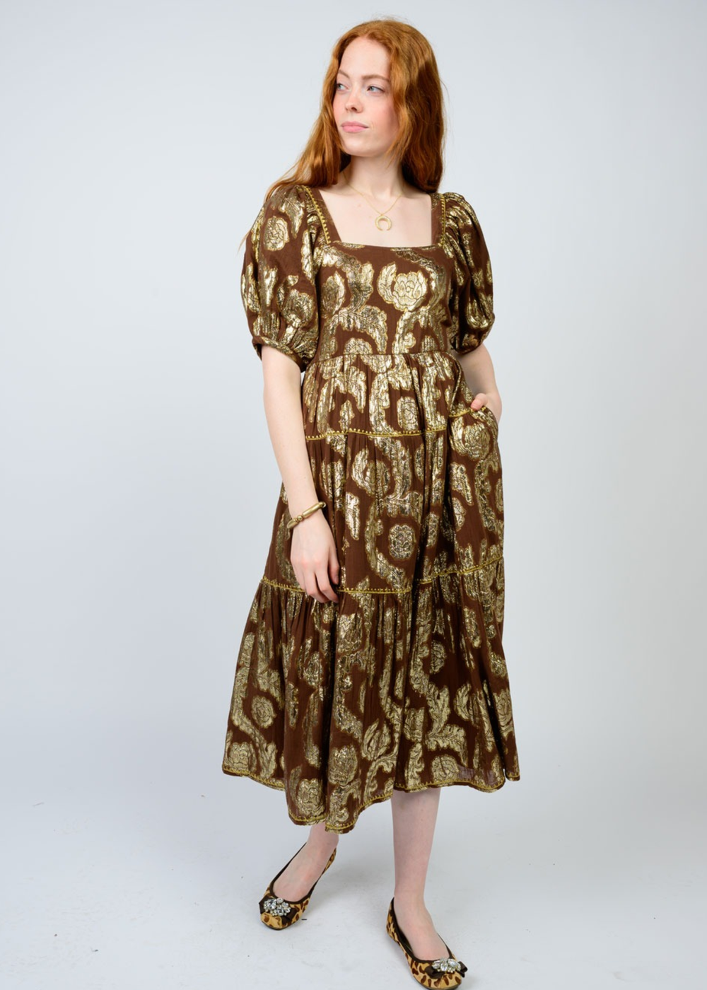 Woman with hand in pocket wearing a brown midi dress with a gold floral pattern