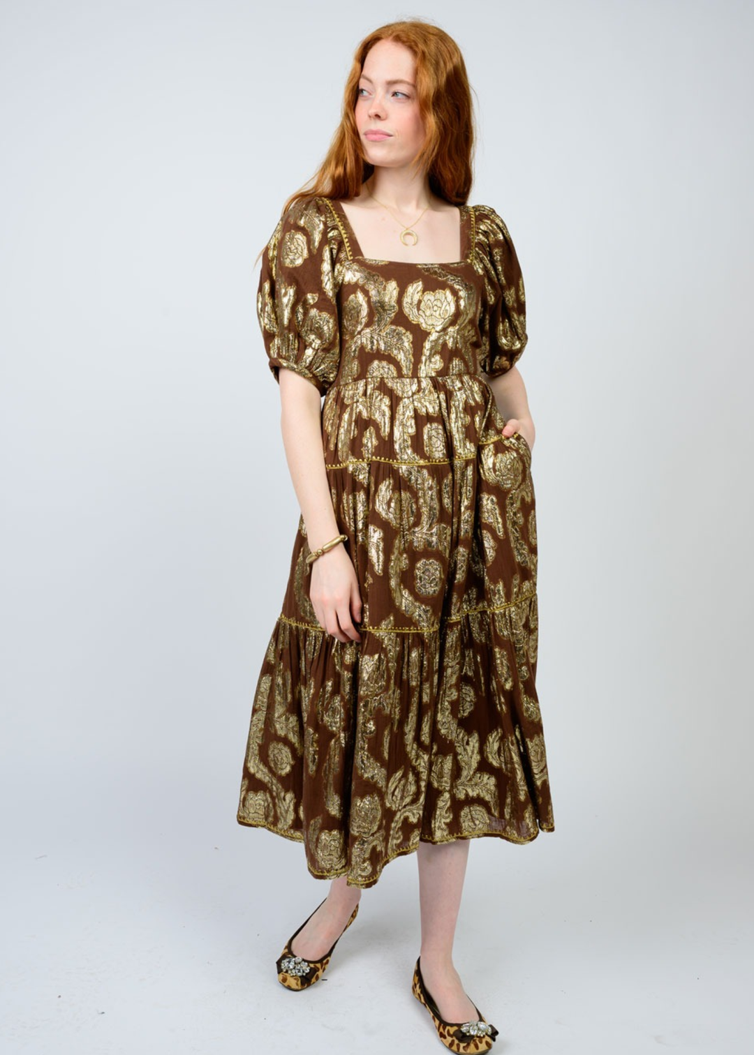 Woman with hand in pocket wearing a brown midi dress with a gold floral pattern